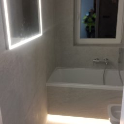 Usługi Ogólnobudowlane Paweł Soboń - Nowoczesna łazienka z szarymi płytkami, podświetlanym lustrem i podłogą, widoczna wanna i fragment toalety, osoba w odblaskowej kamizelce odbija się w oknie.