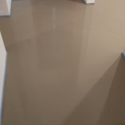 Usługi Ogólnobudowlane Paweł Soboń - Świeżo wylana, samopoziomująca wylewka podłogowa w Głoskowie, przygotowana pod dalsze prace wykończeniowe, z widocznym zabezpieczeniem folią drzwi i listwami piankowymi przy ścianach.