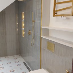 Usługi Ogólnobudowlane Paweł Soboń - Nowoczesna łazienka z szarymi płytkami, złotymi akcentami i wzorzystą podłogą. Kabina prysznicowa typu walk-in, podwieszana toaleta i grzejnik drabinkowy.