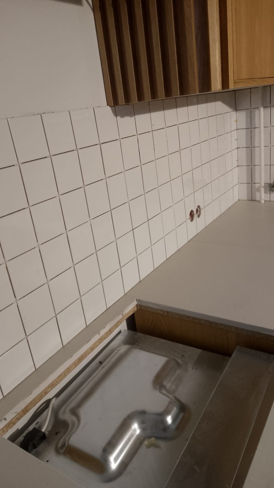 Białe, kwadratowe płytki na ścianie nad blatem kuchennym z wycięciem na zlew i widoczną instalacją hydrauliczną pod spodem.