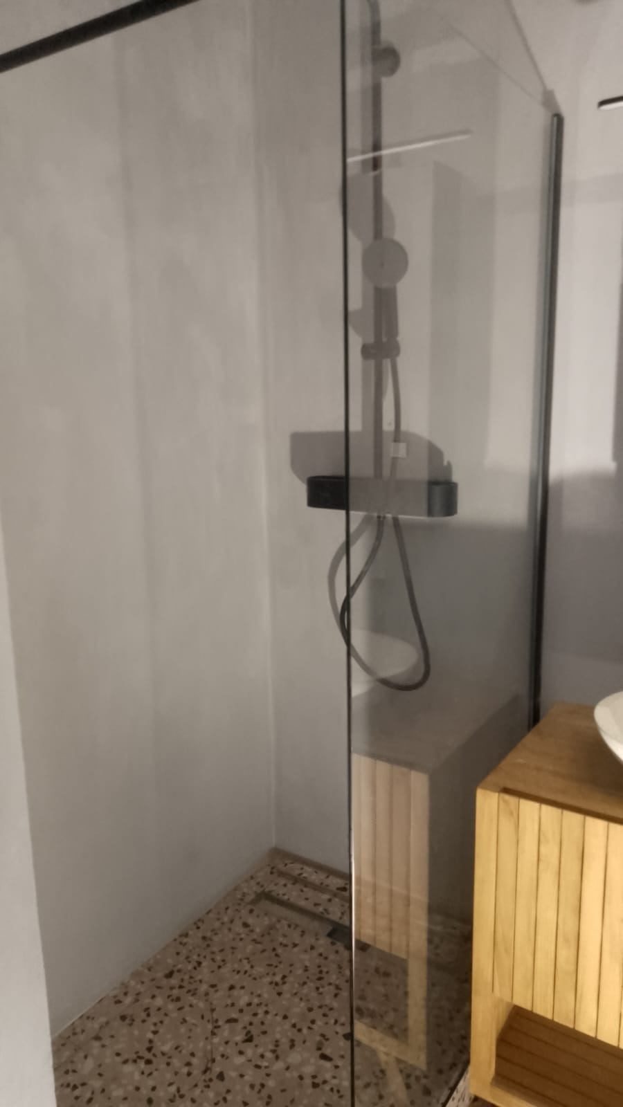 Nowoczesna kabina prysznicowa z deszczownicą i odpływem liniowym w podłodze wyłożonej mozaiką, obok szafka łazienkowa z drewna.