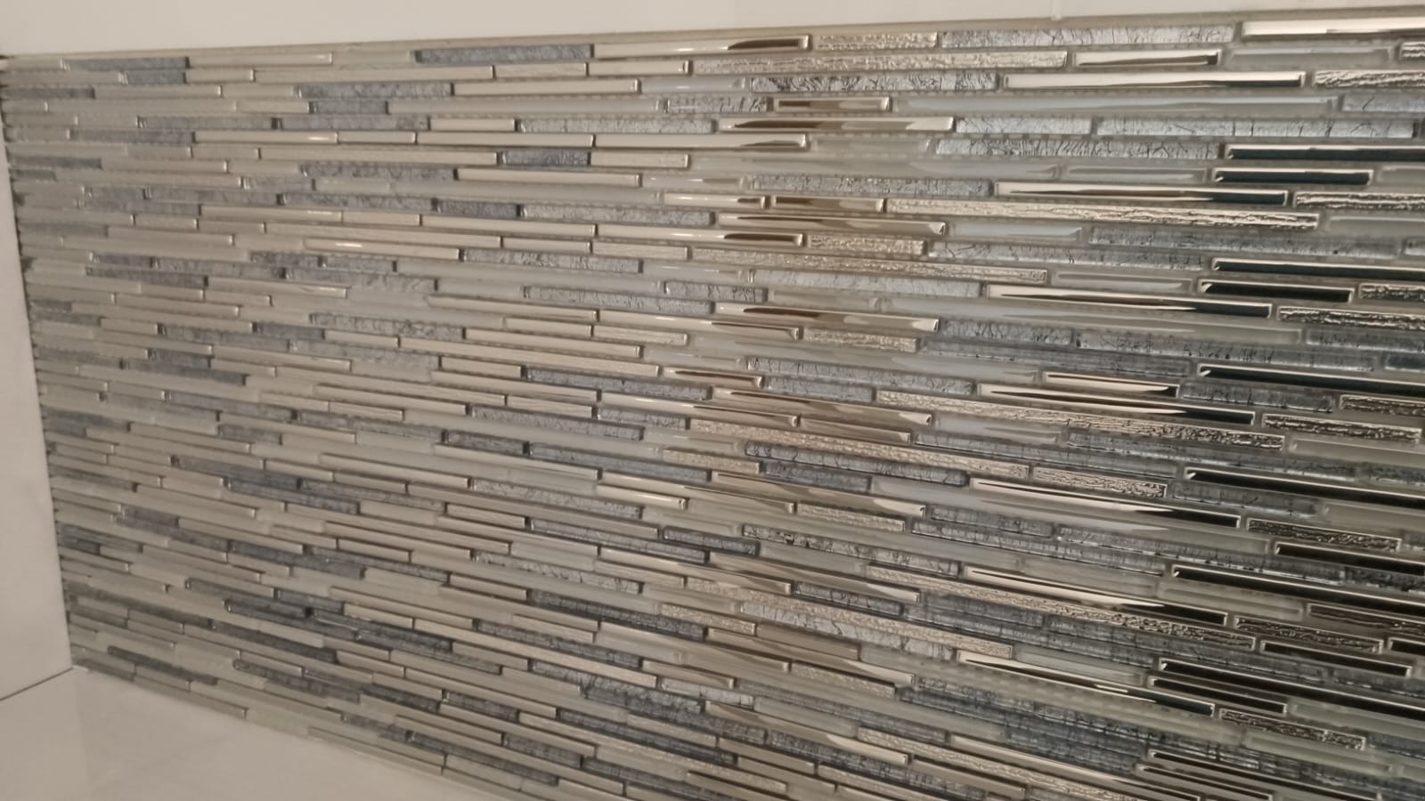 Mozaika ścienna z wąskich, poziomych płytek w odcieniach szarości, srebra i bieli, z elementami o metalicznym połysku i zróżnicowanej fakturze, ułożona na ścianie w łazience.