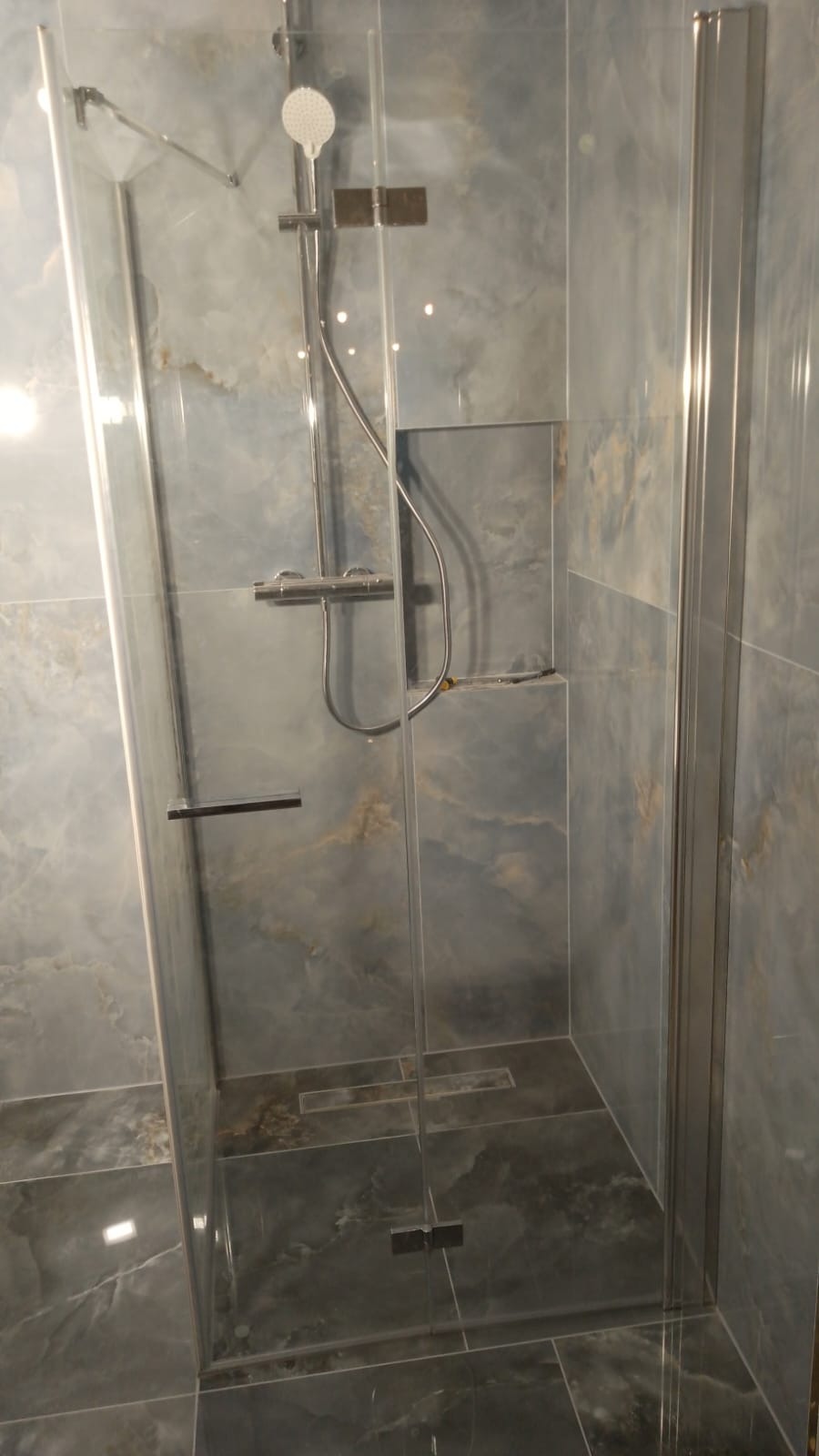 Szklana kabina prysznicowa z deszczownicą i odpływem liniowym w wykończonej łazience z płytkami imitującymi marmur.