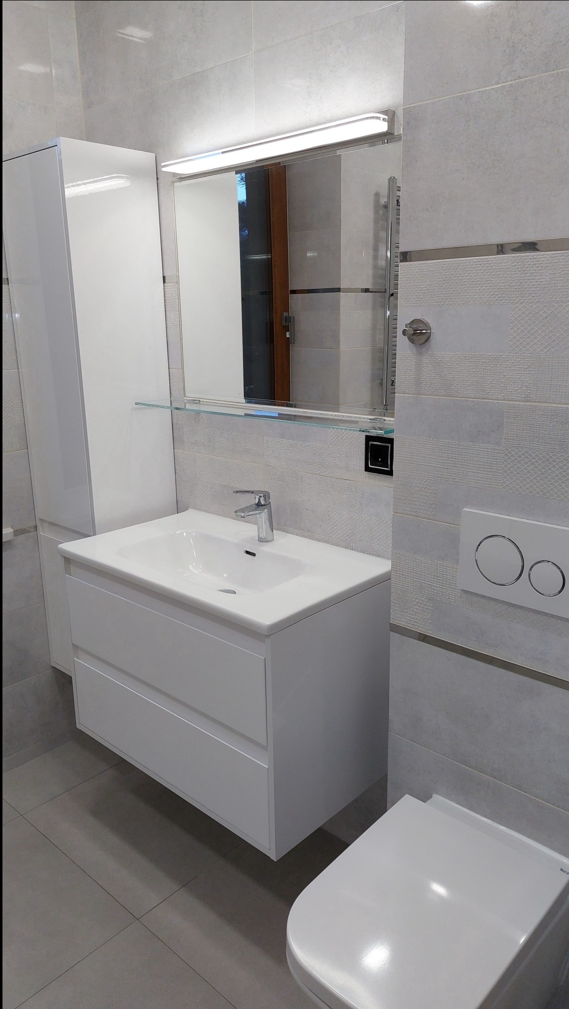 Nowoczesna łazienka z białymi szafkami pod umywalką, lustrem z oświetleniem LED, szarą glazurą na ścianach i podłodze oraz wiszącą toaletą.