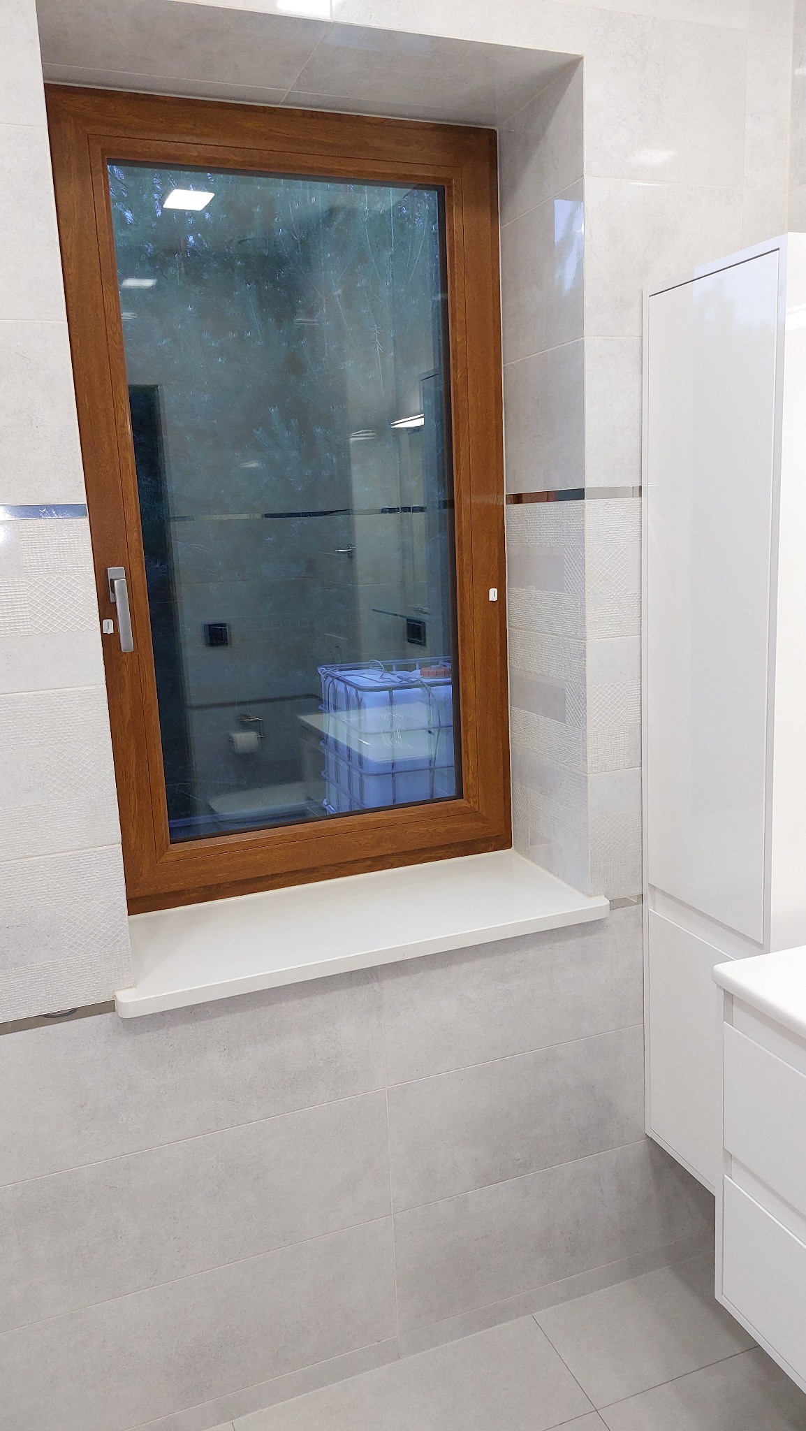 Wykończona łazienka z drewnianym oknem, jasnymi płytkami i białą szafką, widoczne odbicie wnętrza łazienki w szybie okna.