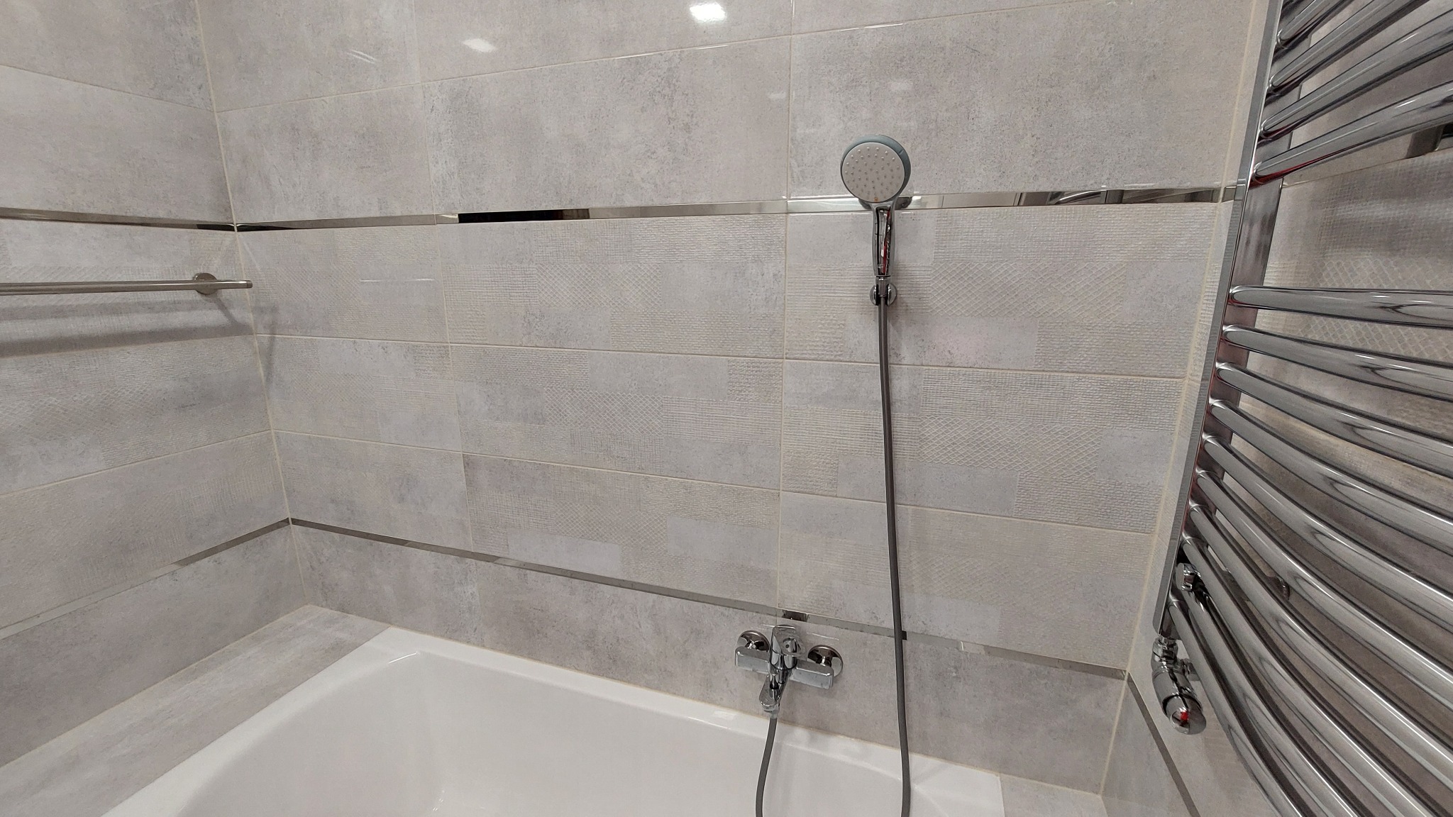 Jasnoszara łazienka z wanną, chromowaną armaturą, grzejnikiem drabinkowym i delikatnym wzorem na płytkach.