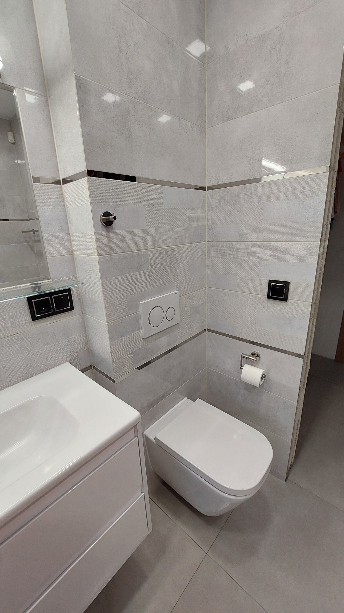 Nowoczesna toaleta podwieszana z białym panelem spłukującym na ścianie z szarych płytek, obok umywalka z szafką w minimalistycznym stylu.