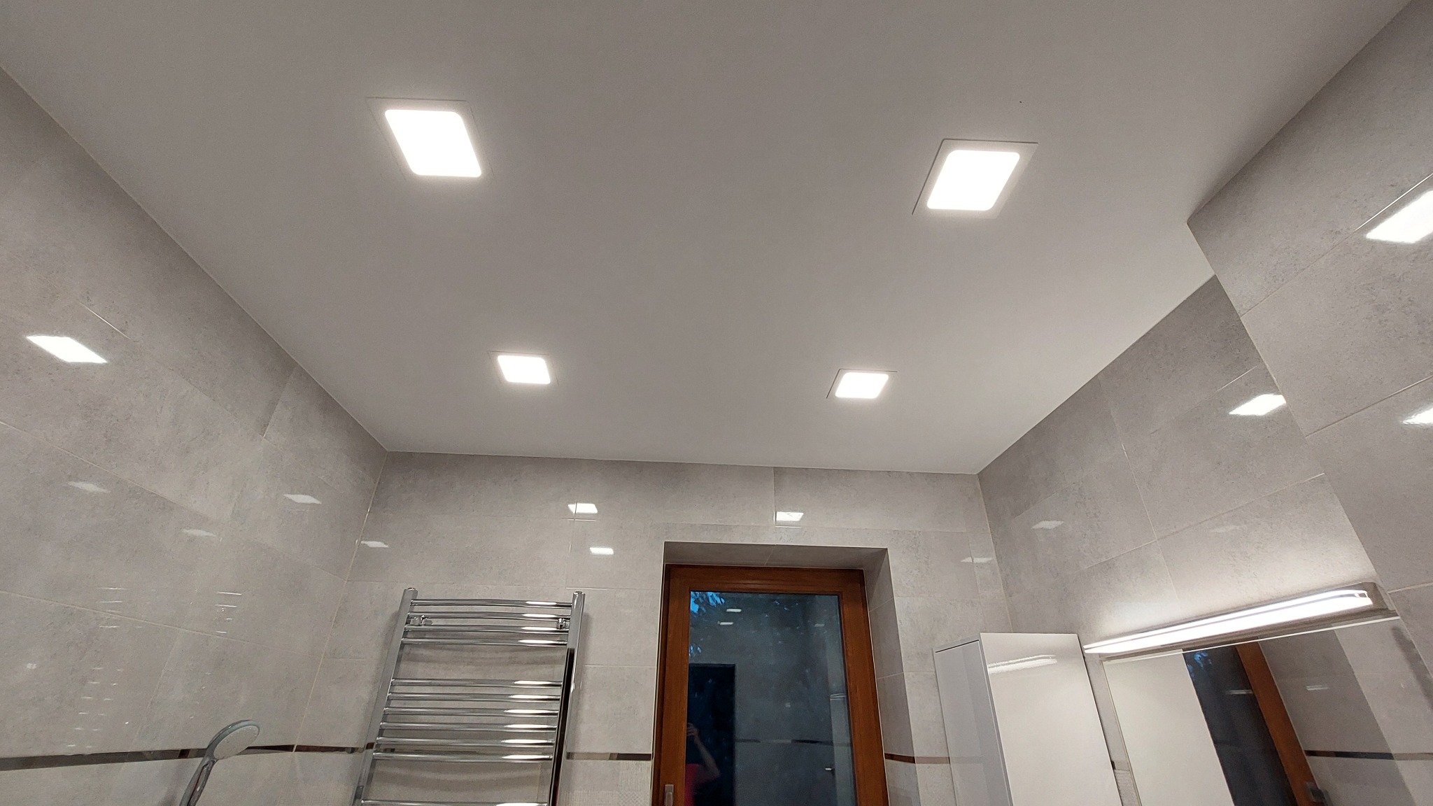 Wykończona łazienka z szarymi płytkami, podwieszanym sufitem z czterema kwadratowymi lampami LED, grzejnikiem drabinkowym i szafką pod umywalką.