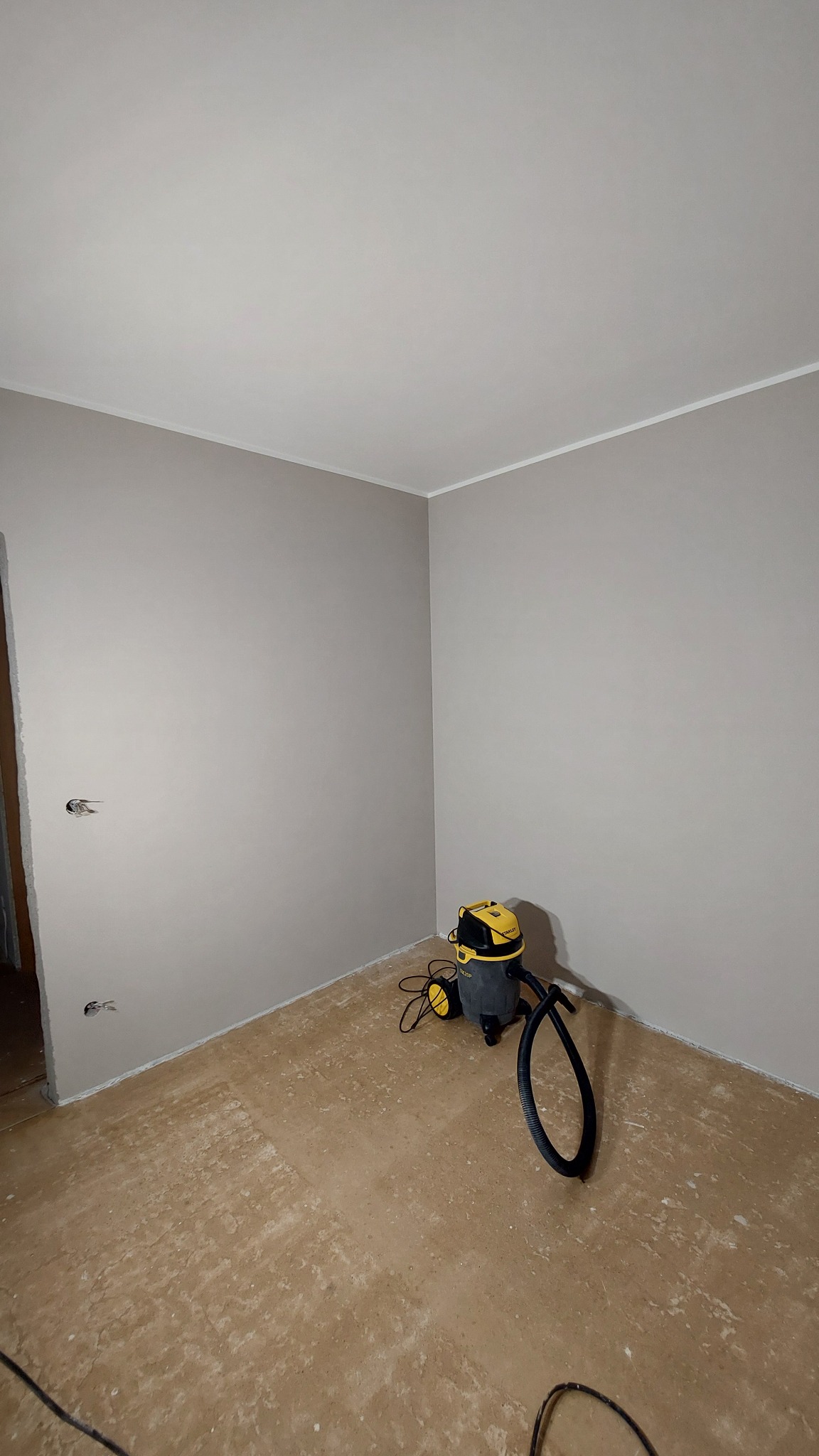 Świeżo pomalowane na szaro ściany w rogu pokoju, widoczne gniazdka elektryczne bez osłon, przygotowana podłoga i odkurzacz przemysłowy Stanley SXVC20P.