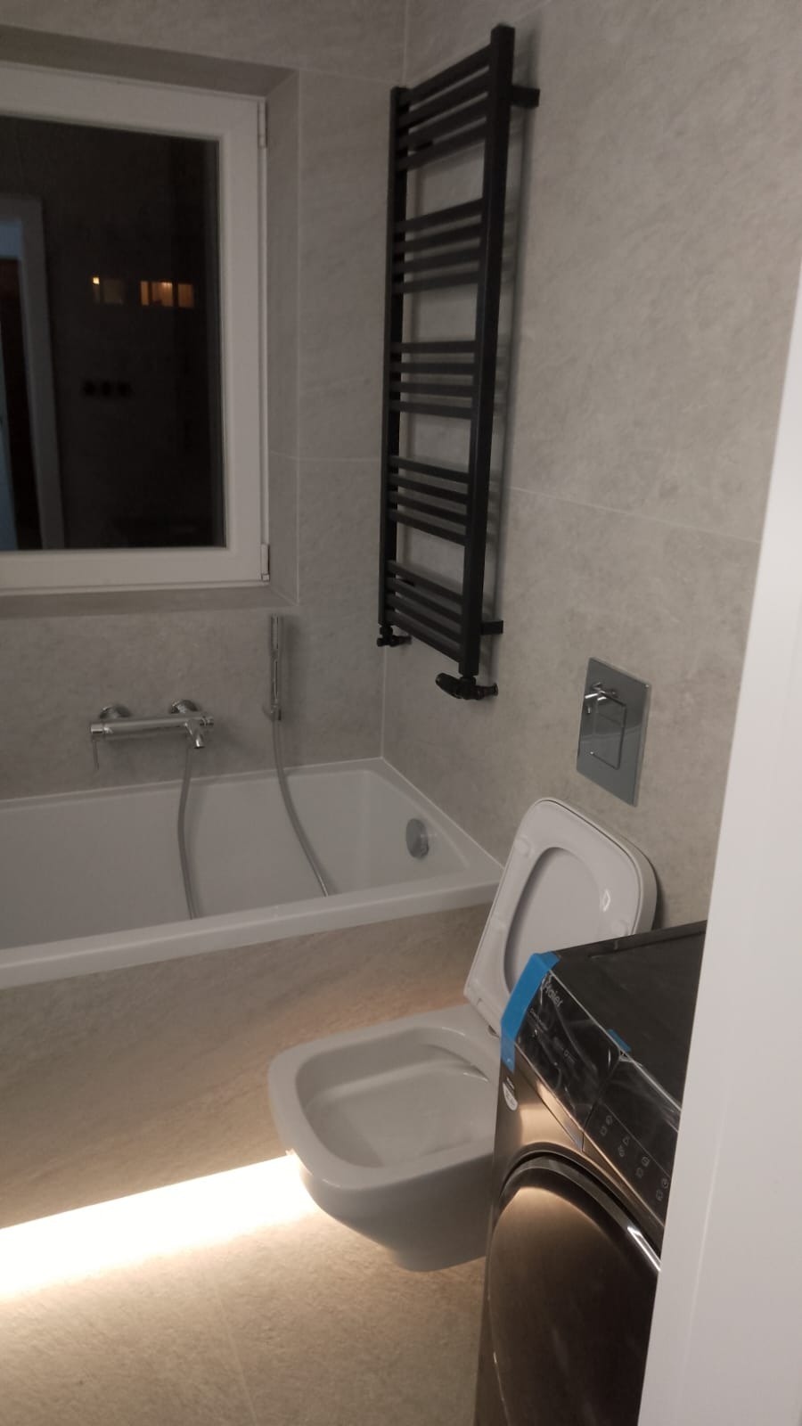 Jasna łazienka z wanną, toaletą podwieszaną i grzejnikiem drabinkowym w kolorze czarnym, widoczna pralka z folią ochronną.