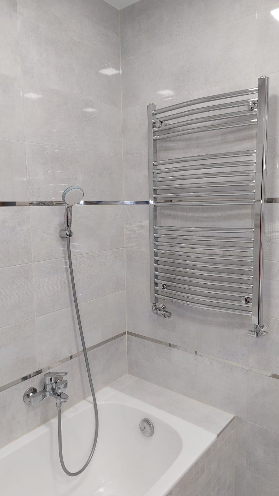 Nowoczesna łazienka z jasnymi, kwadratowymi płytkami, chromowaną armaturą i grzejnikiem drabinkowym, wykończona srebrną listwą dekoracyjną.