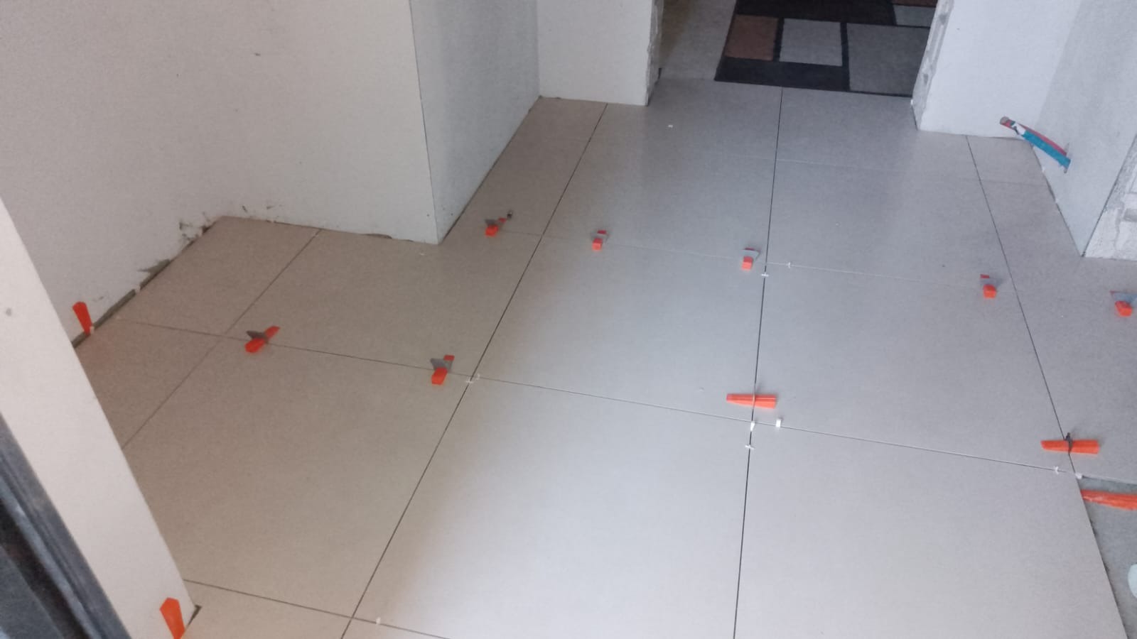 Świeżo ułożone, jasnoszare płytki podłogowe z pomarańczowymi klipsami poziomującymi i krzyżykami dystansowymi, widoczne ślady zaprawy na ścianie przy podłodze.