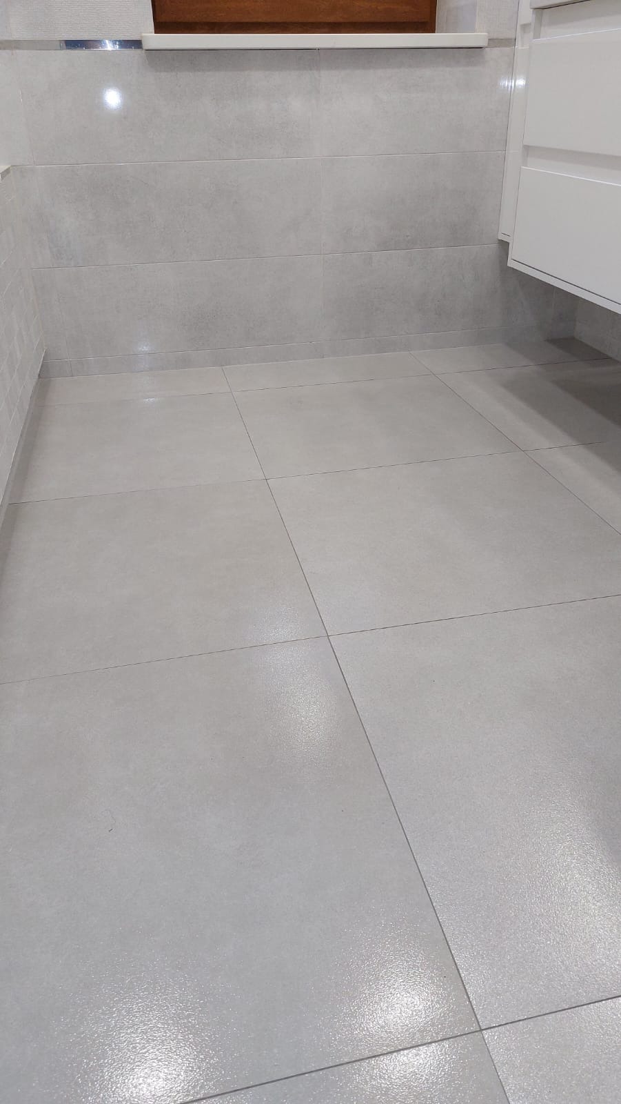 Jasnoszara, matowa podłoga z płytek w formacie kwadratu, ułożona w regularną siatkę w wykończonej łazience, z fragmentem białej szafki pod umywalkę po prawej stronie i listwą przypodłogową w tle.