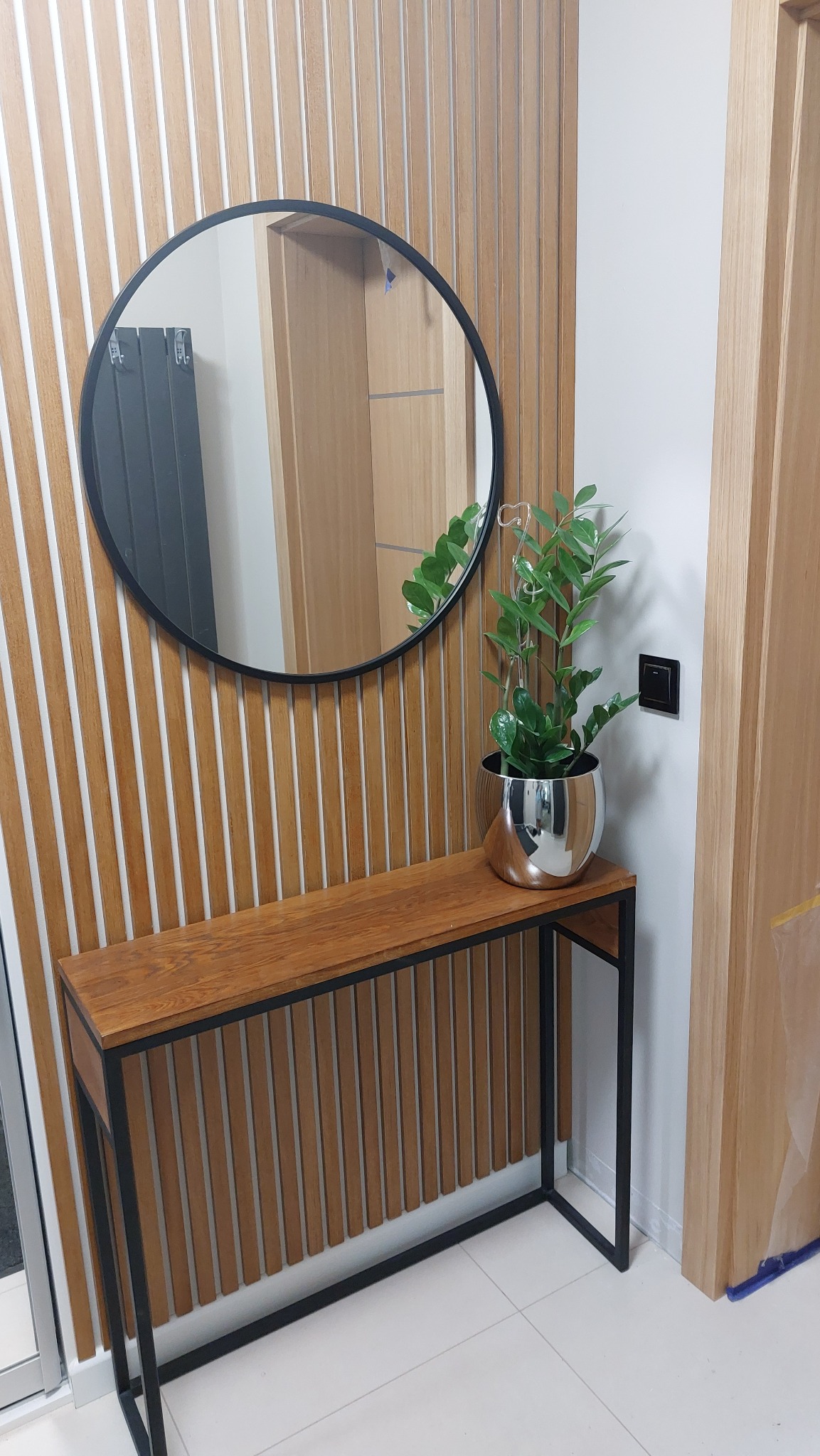 Nowoczesny przedpokój: drewniana konsola z czarnymi nogami, okrągłe lustro i roślina w srebrnej doniczce na tle ściany z drewnianych lameli.