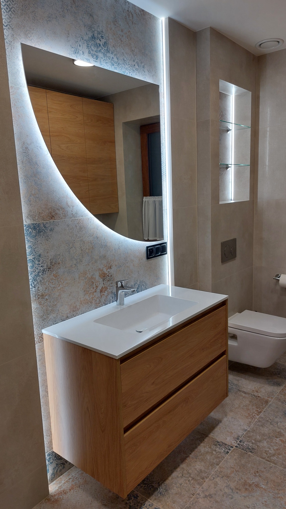 Nowoczesna łazienka z podświetlanym lustrem o nietypowym kształcie, drewnianą szafką pod umywalką i płytkami w stylu vintage. Widoczna toaleta i wnęka z półkami.