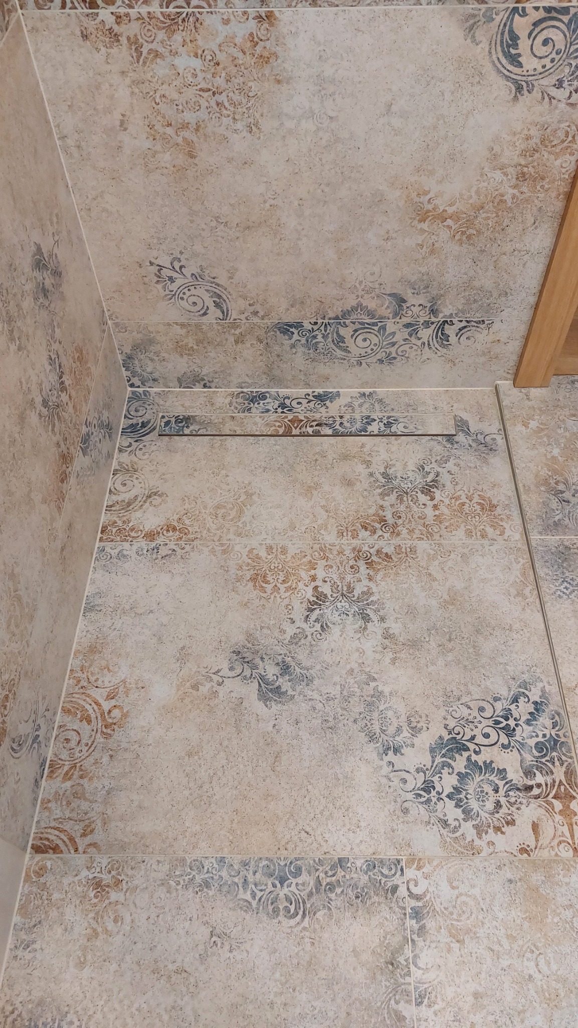 Beżowe płytki z ornamentami na ścianie i podłodze, ułożone w rogu pomieszczenia. Widoczny fragment drewnianej listwy. Dekoracyjne wykończenie wnętrza.