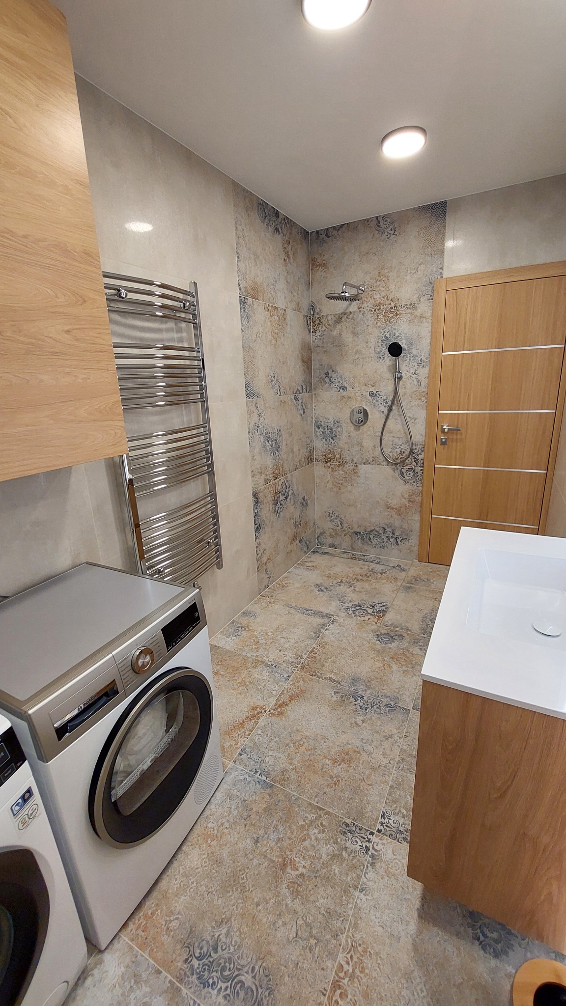 Wykończona łazienka z kabiną prysznicową typu walk-in, pralką i grzejnikiem drabinkowym. Płytki w odcieniach beżu i błękitu z wzorem. Drewniane akcenty.