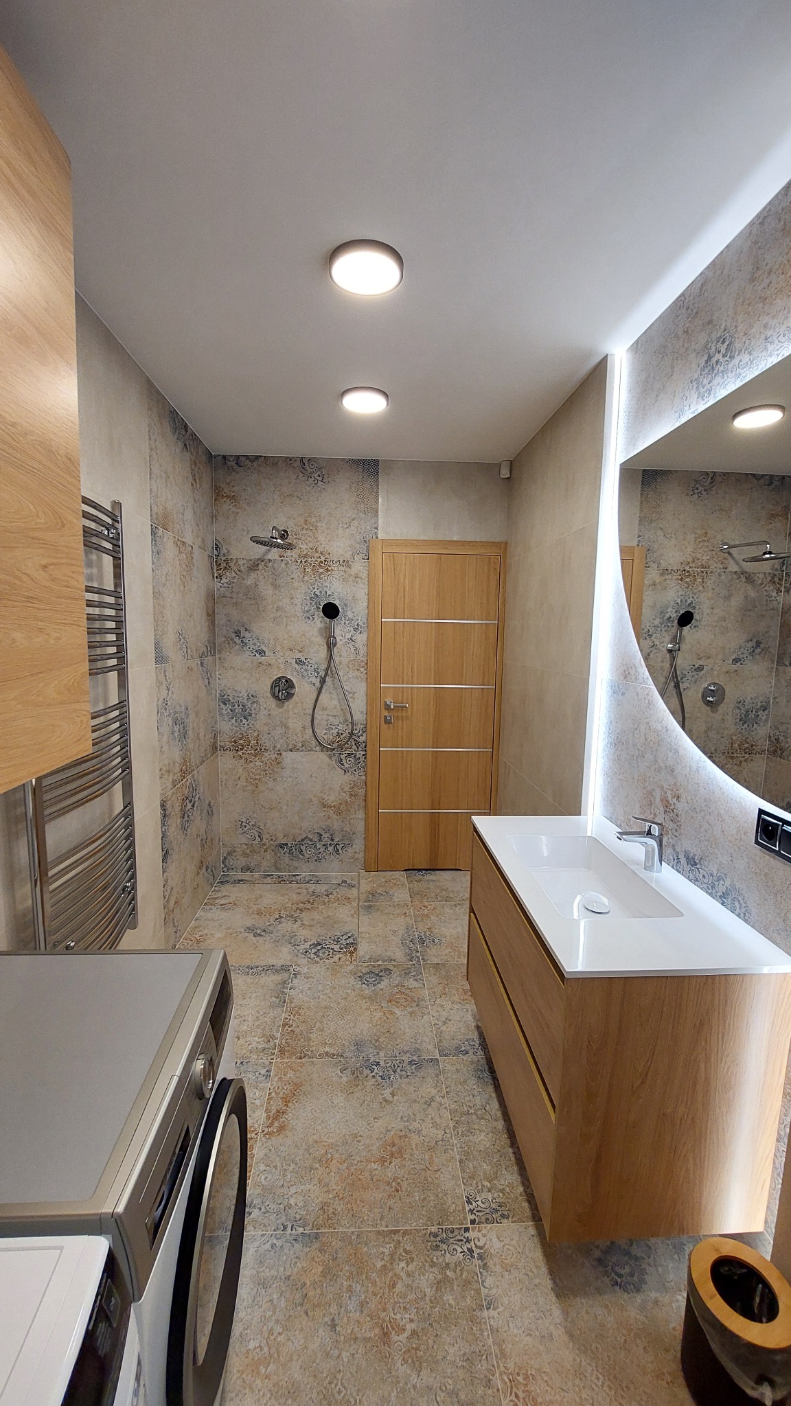 Nowoczesna łazienka z kabiną prysznicową typu walk-in, umywalką z szafką z jasnego drewna i dużym lustrem z podświetleniem LED. Na podłodze i ścianach wzorzyste płytki.