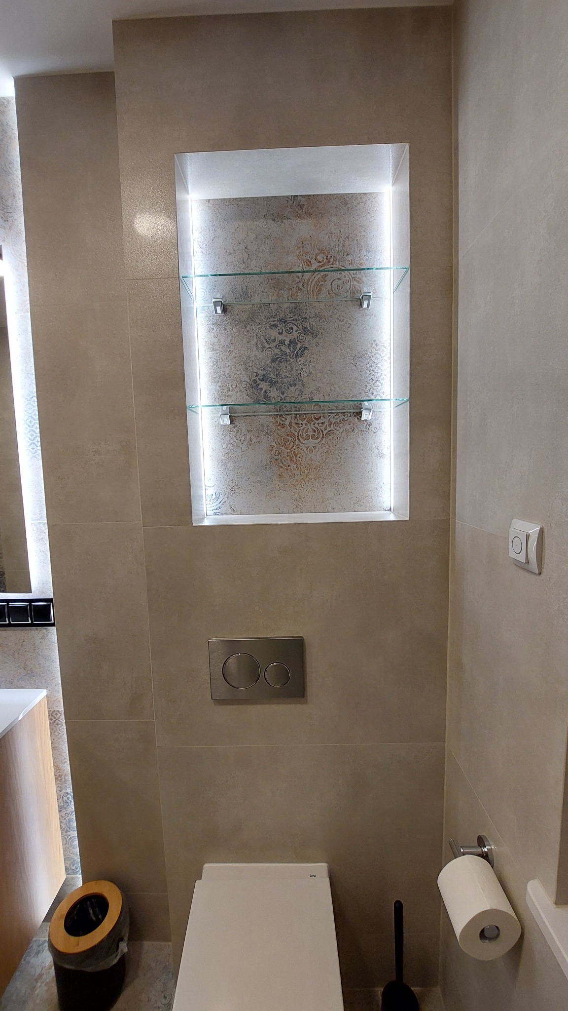 Beżowa łazienka z podświetlaną wnęką z półkami, wiszącą toaletą i akcesoriami. Minimalistyczny design, płytki imitujące kamień, nowoczesne oświetlenie.