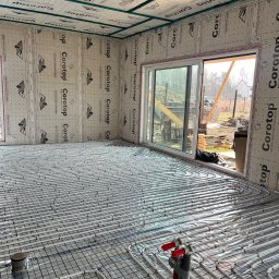 DOSKONAŁY DOM SPÓŁKA Z OGRANICZONĄ ODPOWIEDZIALNOŚCIĄ - Instalacja ogrzewania podłogowego w domu szkieletowym, widoczne rury na folii z nadrukiem, okno przesuwne z widokiem na plac budowy.