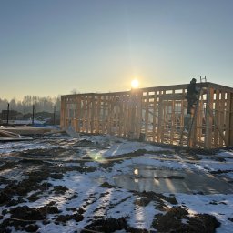 DOSKONAŁY DOM SPÓŁKA Z OGRANICZONĄ ODPOWIEDZIALNOŚCIĄ - Konstrukcja domu szkieletowego w zimowym krajobrazie, z pracownikiem na drabinie i promieniami słońca w tle. Widoczne elementy drewniane i zasnieżona ziemia.