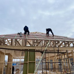 DOSKONAŁY DOM SPÓŁKA Z OGRANICZONĄ ODPOWIEDZIALNOŚCIĄ - Budowa domu szkieletowego: Dwóch monterów na dachu z czerwonym pokryciem, drewniana konstrukcja w surowym stanie, drabiny i narzędzia w tle, pochmurne niebo.