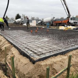 DOSKONAŁY DOM SPÓŁKA Z OGRANICZONĄ ODPOWIEDZIALNOŚCIĄ - Wylewka fundamentowa zbrojona siatką stalową, widoczne szalunki i ekipa w trakcie pracy. W tle betoniarki i teren budowy w pochmurny dzień.