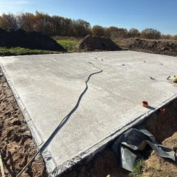 DOSKONAŁY DOM SPÓŁKA Z OGRANICZONĄ ODPOWIEDZIALNOŚCIĄ - Wylana płyta fundamentowa pod dom szkieletowy, widok z góry. Na obrzeżach widoczne zabezpieczenia i wykop. W tle drzewa i błękitne niebo.