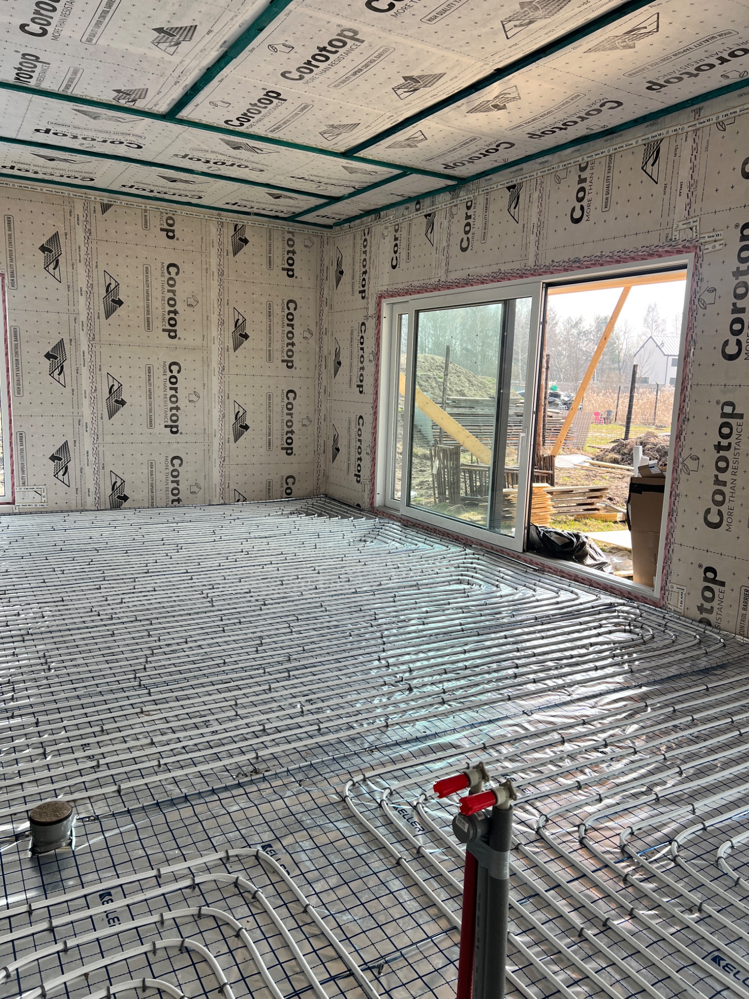 Instalacja ogrzewania podłogowego w domu szkieletowym, widoczne rury na folii z nadrukiem, okno przesuwne z widokiem na plac budowy.