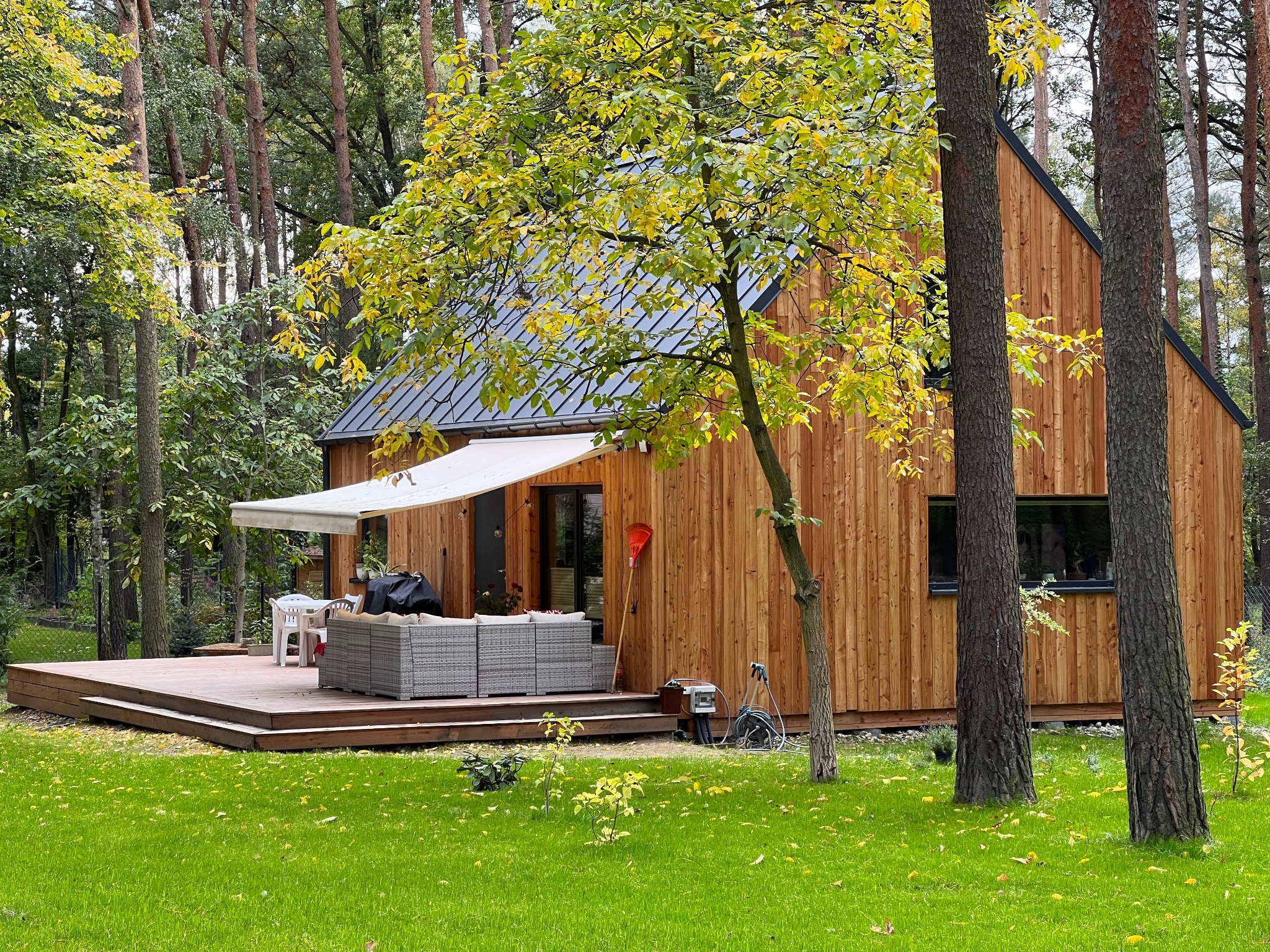 Nowoczesny dom szkieletowy z drewnianą elewacją i tarasem, otoczony zielenią. Widoczny fragment ogrodu z meblami wypoczynkowymi i drzewami.