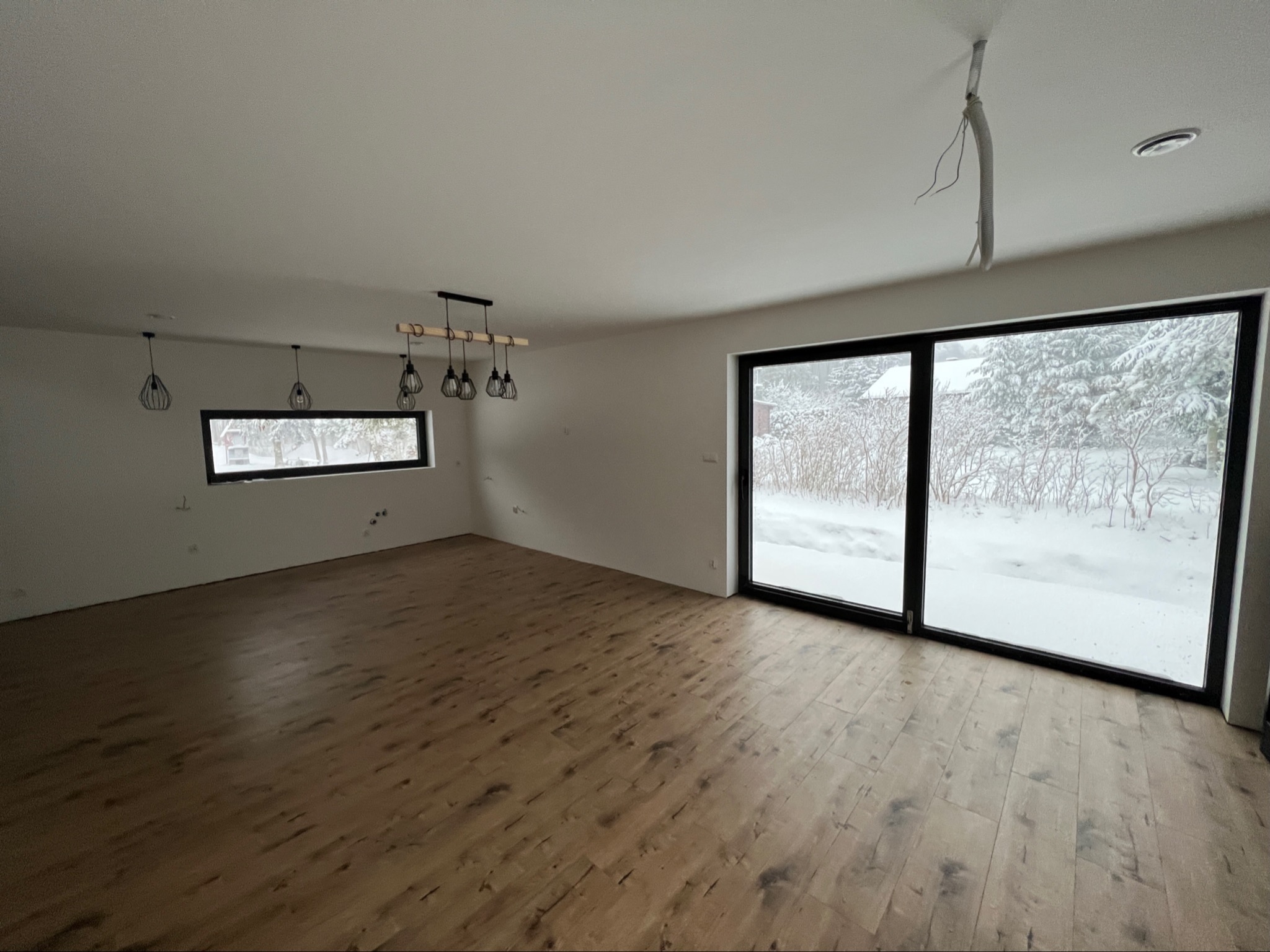 Wnętrze domu szkieletowego z drewnianą podłogą i dużymi oknami z widokiem na zimowy krajobraz. Widoczne lampy i niedokończone instalacje na suficie.