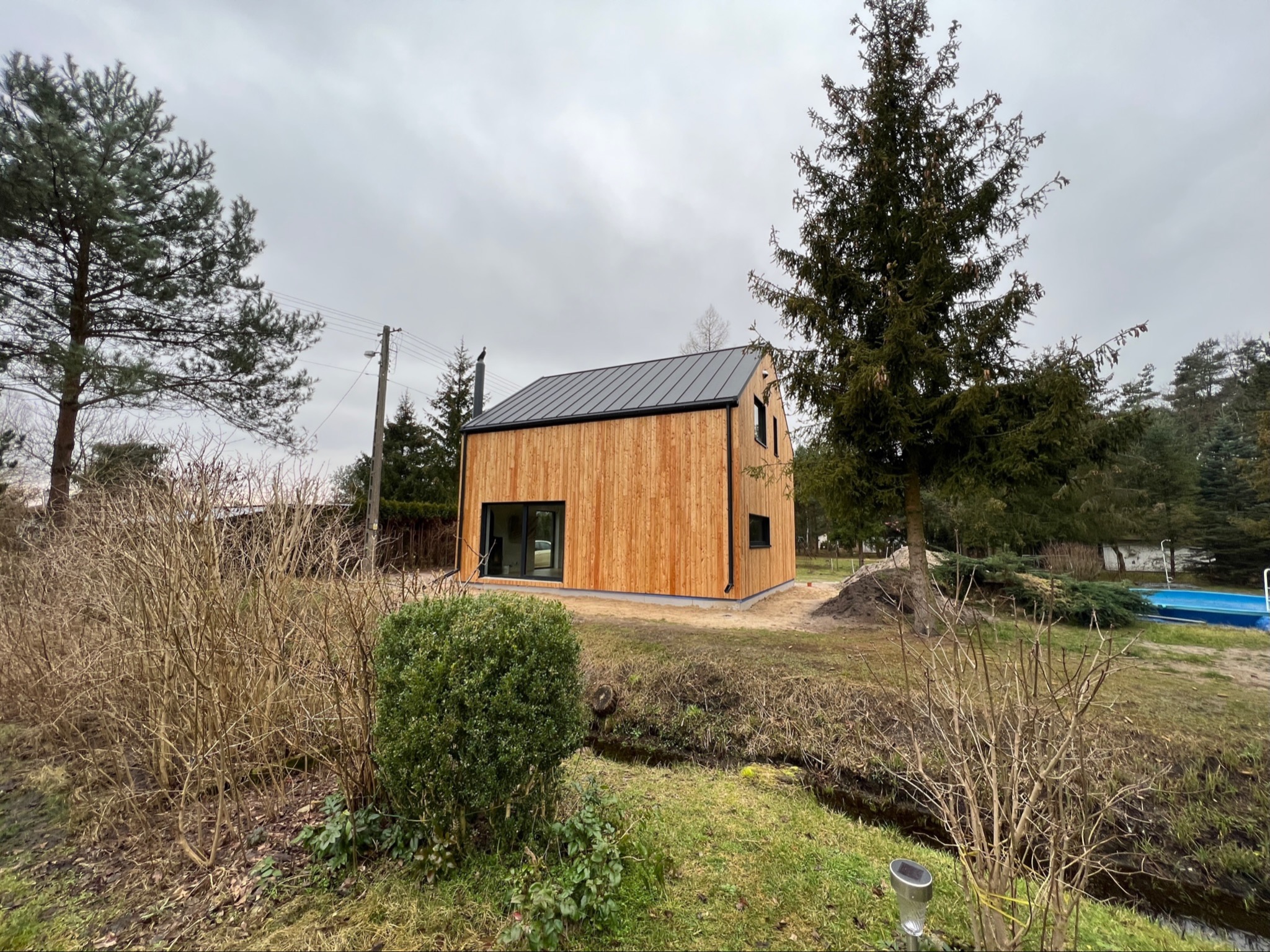 Nowoczesny dom szkieletowy z drewnianą elewacją i czarnym dachem na tle pochmurnego nieba. Widoczny fragment basenu i drzewa iglaste.