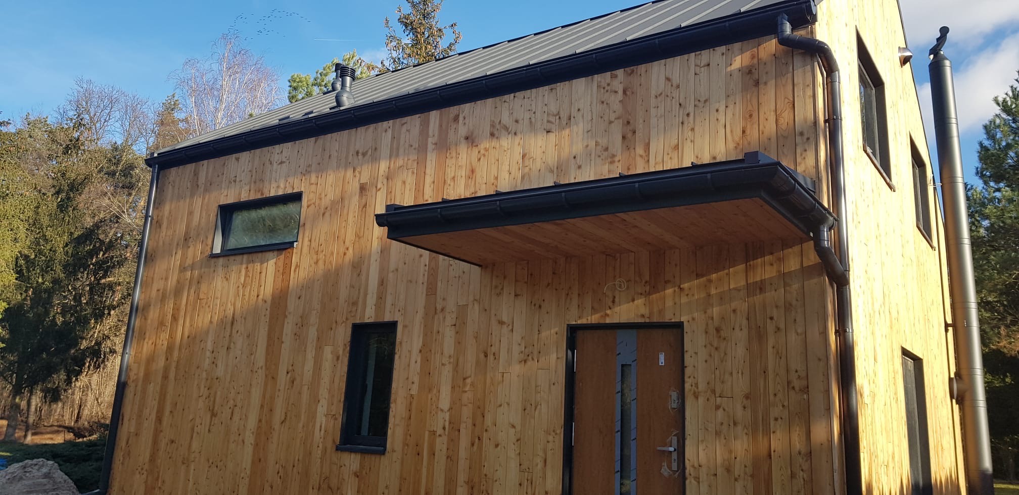 Nowoczesny dom szkieletowy z drewnianą elewacją, czarnymi oknami i orynnowaniem. Widoczny fragment dachu i komin. W tle drzewa i błękitne niebo.