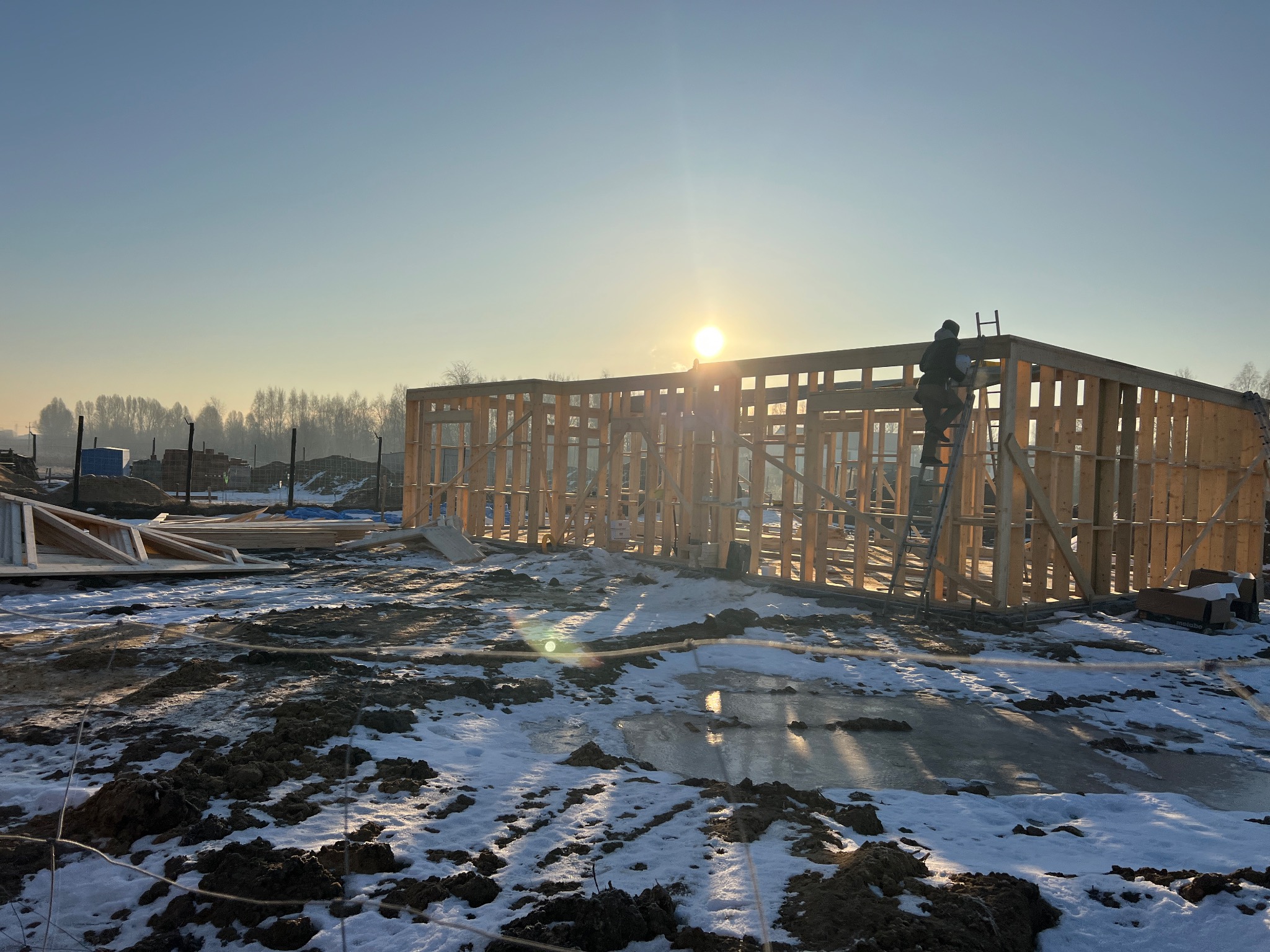 Konstrukcja domu szkieletowego w zimowym krajobrazie, z pracownikiem na drabinie i promieniami słońca w tle. Widoczne elementy drewniane i zasnieżona ziemia.