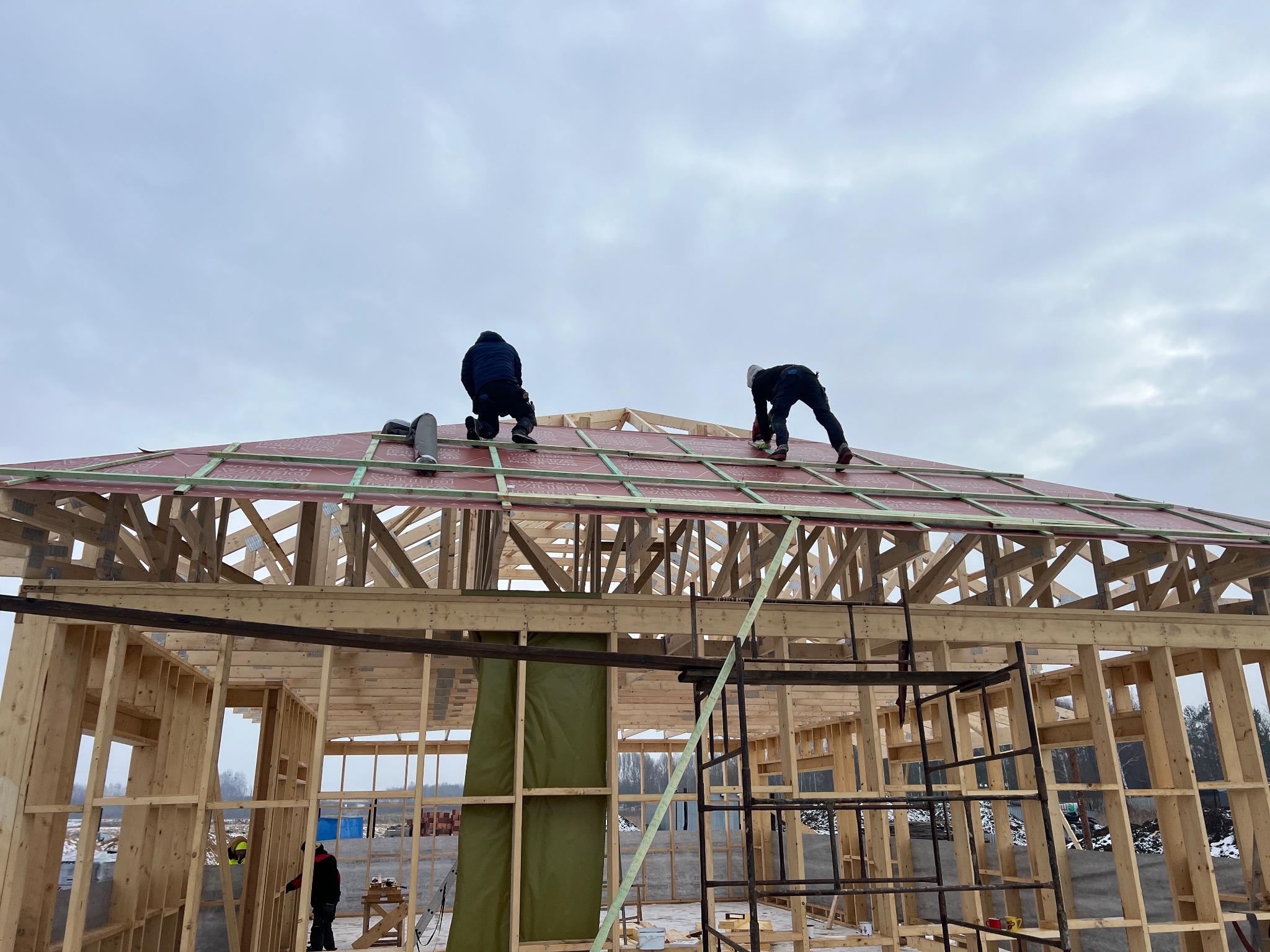 Budowa domu szkieletowego: Dwóch monterów na dachu z czerwonym pokryciem, drewniana konstrukcja w surowym stanie, drabiny i narzędzia w tle, pochmurne niebo.