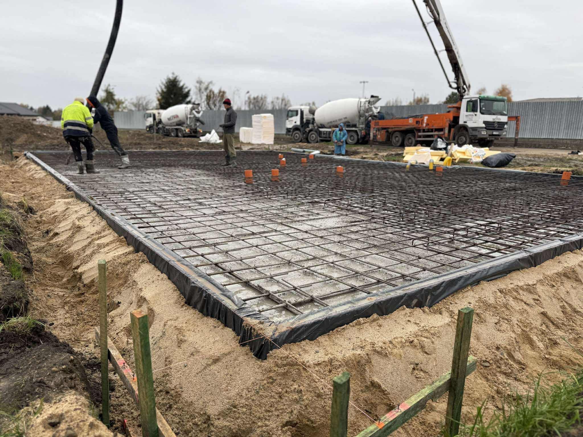 Wylewka fundamentowa zbrojona siatką stalową, widoczne szalunki i ekipa w trakcie pracy. W tle betoniarki i teren budowy w pochmurny dzień.