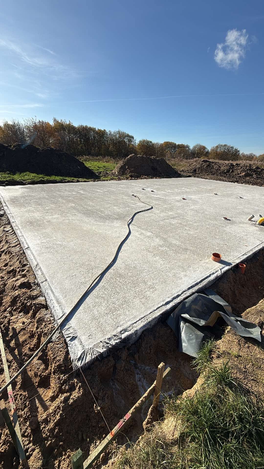 Wylana płyta fundamentowa pod dom szkieletowy, widok z góry. Na obrzeżach widoczne zabezpieczenia i wykop. W tle drzewa i błękitne niebo.