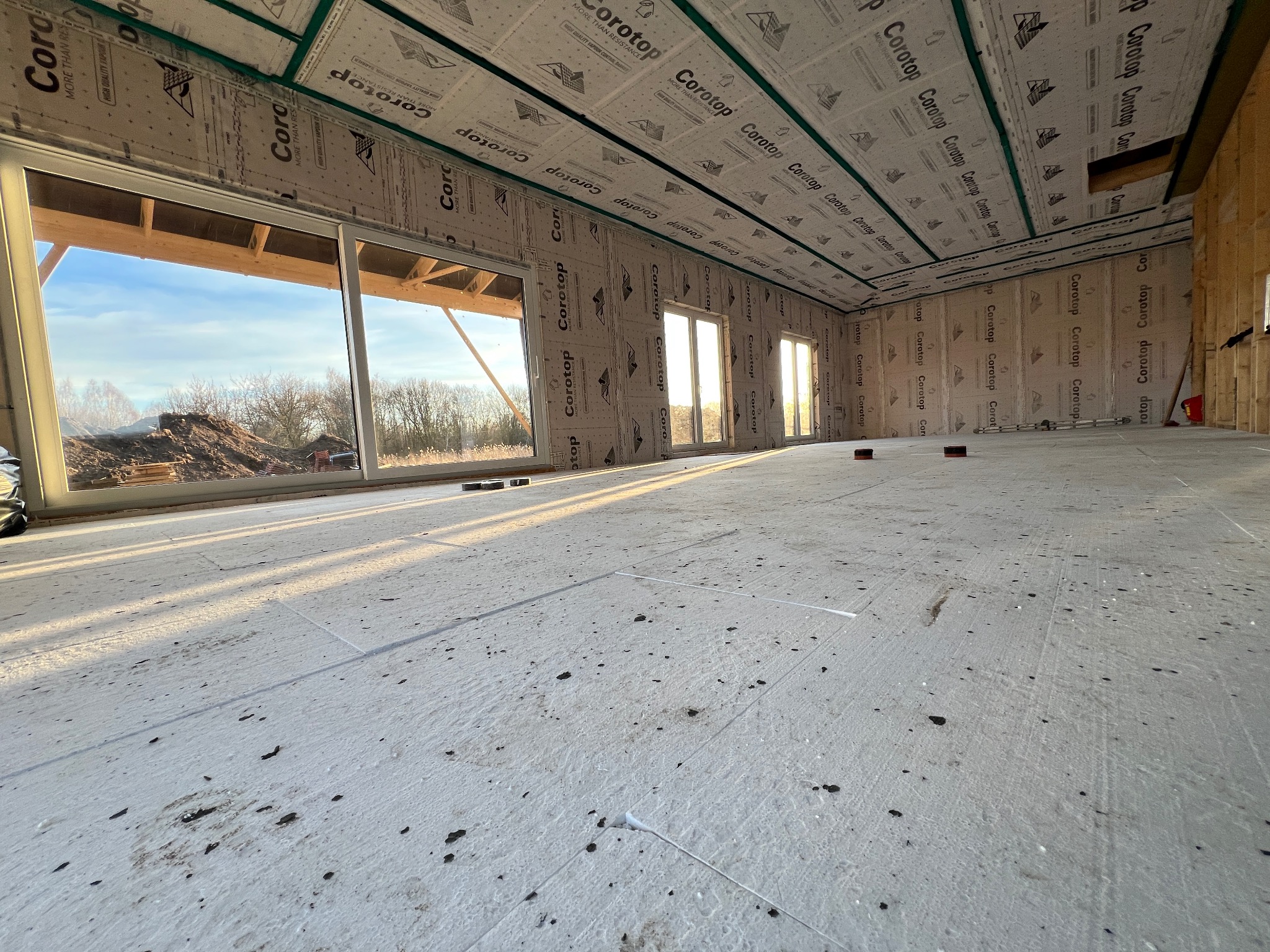 Wnętrze domu szkieletowego w trakcie budowy. Widoczne okna, izolacja ścian i podłogi. Na zewnątrz hałda ziemi. Jasne światło dzienne.