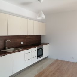 PerfectHome Radosław Matynia - Nowoczesna, jasna kuchnia z białymi, lakierowanymi szafkami i ciemnobrązowym blatem oraz panelem ściennym. Widoczny zlew z szarą baterią, płyta grzewcza i piekarnik w zabudowie. Na podłodze...
