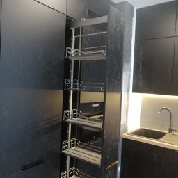 PerfectHome Radosław Matynia - Ciemna kuchnia z wysuwaną szafką cargo z metalowymi półkami, widoczny fragment zlewu i oświetlenie LED pod szafkami.