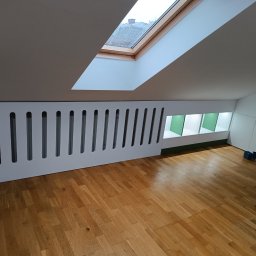 PerfectHome Radosław Matynia - Adaptacja poddasza: Biała zabudowa meblowa z ażurowym frontem w skosie, drewniana podłoga parkietowa, okno dachowe, narzędzia na podłodze wskazują na trwające prace wykończeniowe.