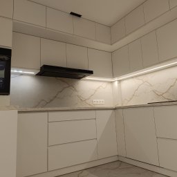 PerfectHome Radosław Matynia - Nowoczesna, minimalistyczna kuchnia z białymi, gładkimi frontami szafek, marmurowym blatem i podświetleniem LED pod szafkami wiszącymi, widoczny piekarnik marki Beko i czarny okap