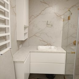 PerfectHome Radosław Matynia - Nowoczesna łazienka z białymi meblami z szufladami i szafką wiszącą, marmurowe płytki na ścianach i podłodze, szklany panel prysznicowy z złotymi okuciami, widoczne opakowanie lampy Zumaline.