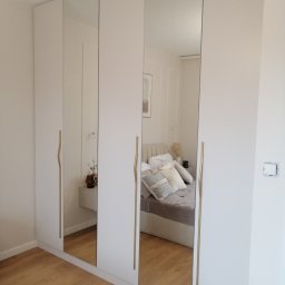 PerfectHome Radosław Matynia - Biała szafa wnękowa z lustrzanymi drzwiami, widoczne złote uchwyty i odbicie sypialni. Minimalistyczny design, jasne wnętrze.