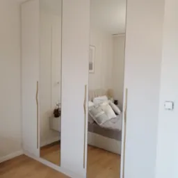 Biała szafa wnękowa z lustrzanymi drzwiami, widoczne złote uchwyty i odbicie sypialni. Minimalistyczny design, jasne wnętrze.