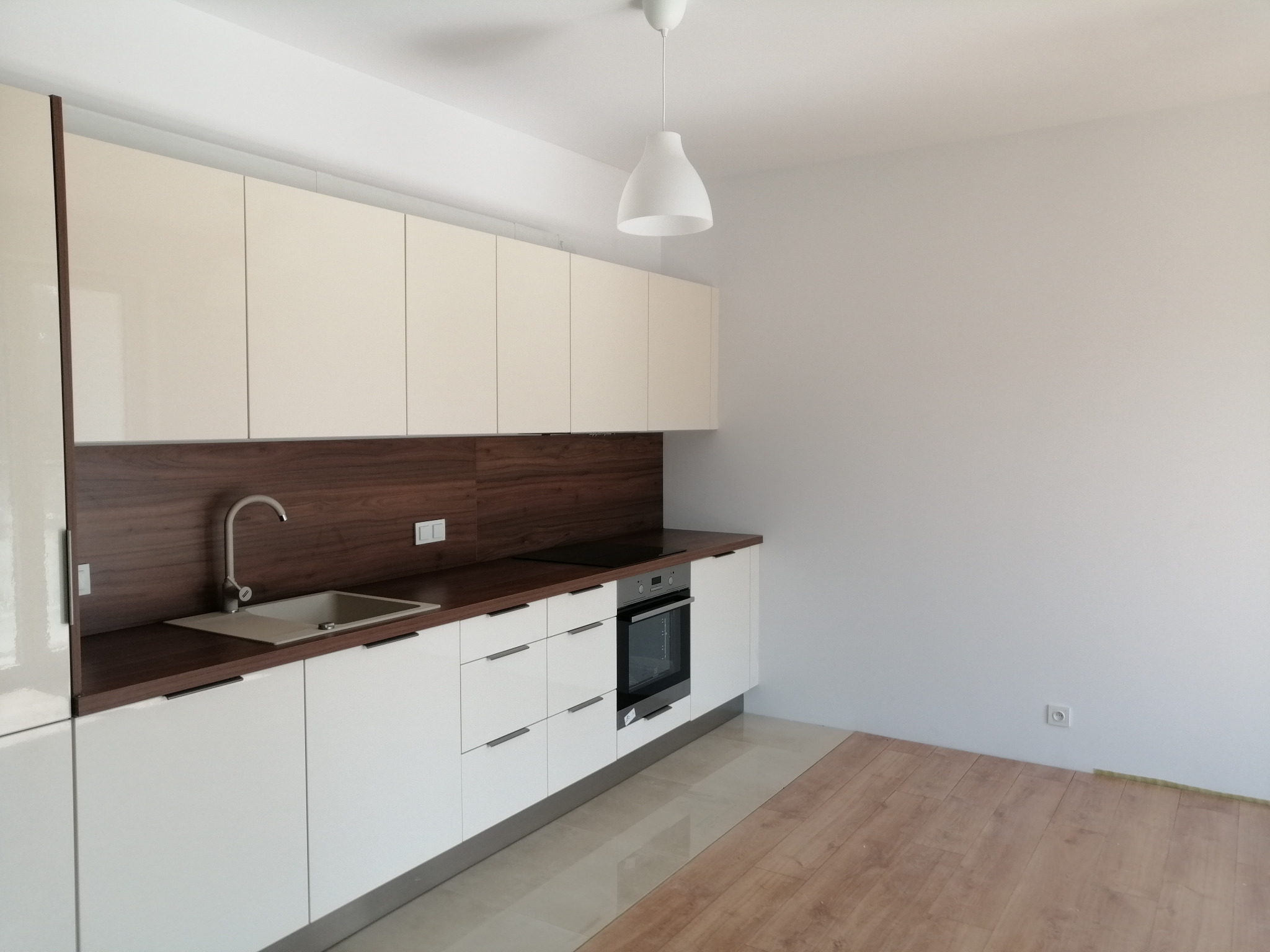 Nowoczesna, jasna kuchnia z białymi, lakierowanymi szafkami i ciemnobrązowym blatem oraz panelem ściennym. Widoczny zlew z szarą baterią, płyta grzewcza i piekarnik w zabudowie. Na podłodze...