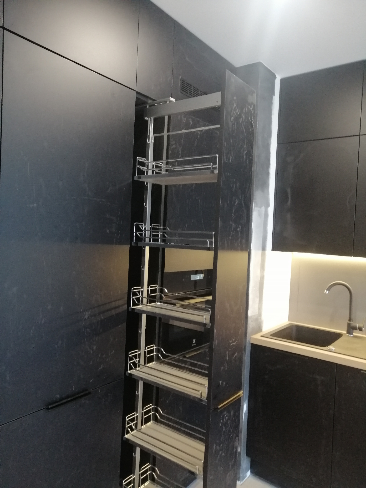 Ciemna kuchnia z wysuwaną szafką cargo z metalowymi półkami, widoczny fragment zlewu i oświetlenie LED pod szafkami.
