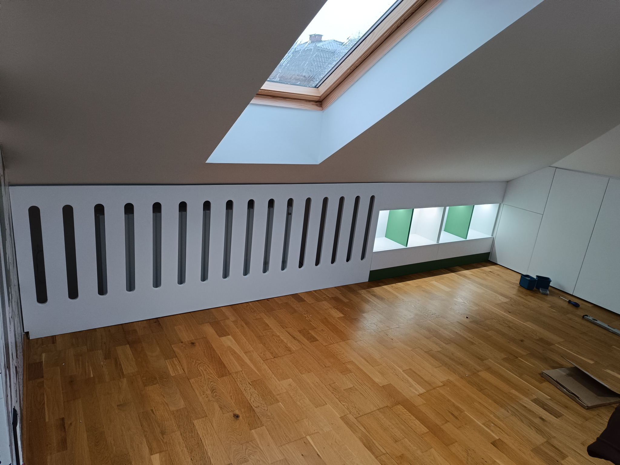 Adaptacja poddasza: Biała zabudowa meblowa z ażurowym frontem w skosie, drewniana podłoga parkietowa, okno dachowe, narzędzia na podłodze wskazują na trwające prace wykończeniowe.