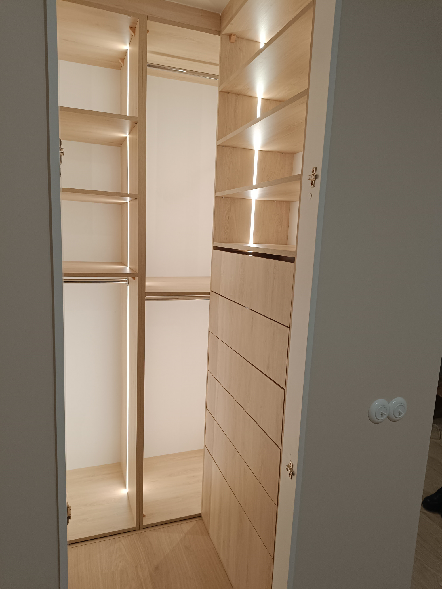 Jasna, drewniana garderoba z oświetleniem LED wewnątrz, widoczne półki, drążek na ubrania i szuflady, nowoczesny design.