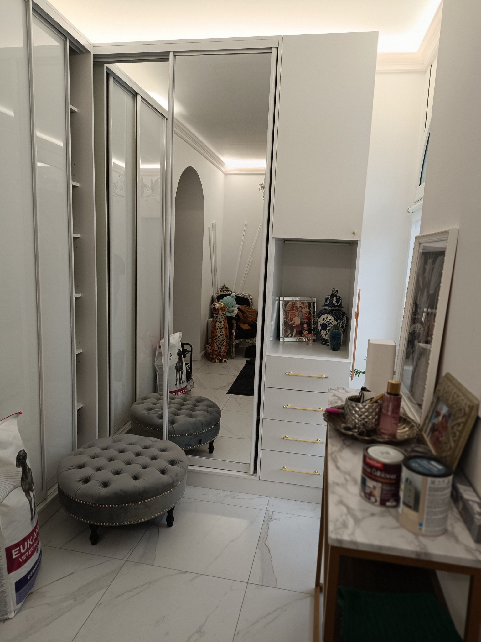 Biała szafa z przesuwnymi, lustrzanymi drzwiami, szary puf i marmurowy stolik z dekoracjami na tle marmurowej podłogi.